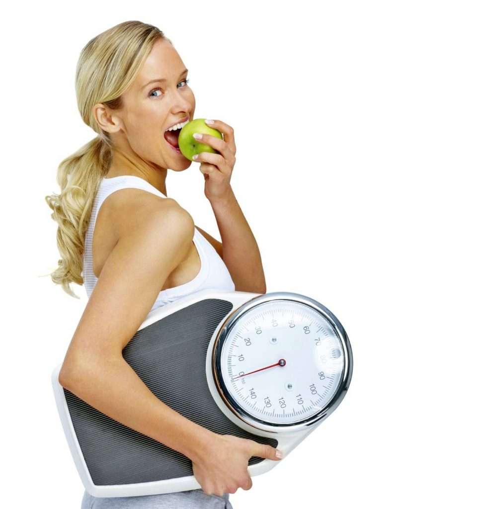 Young woman eating apple and carrying a weight scale over white background