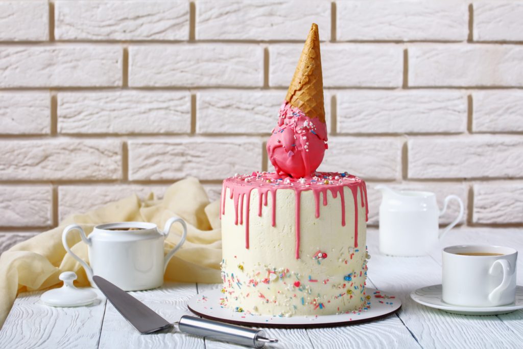 ALT: tort lodowy, polany różową polewą, udekorowany lodem w rożku, w tle łopatka do nakładania ciasta i ceramiczne naczynia